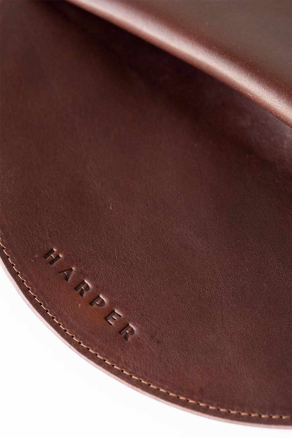 A circular dark brown leather clutch rests against a white background. The focus is on the embossed "Harper" logo and the stitching.