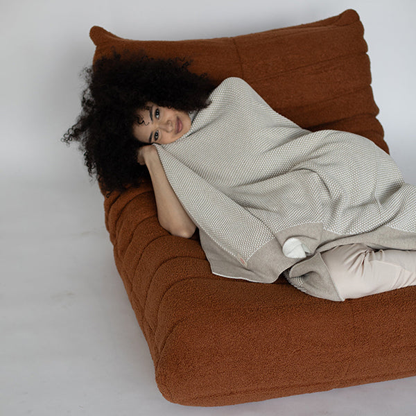 Person wrapped in a soft cotton beige blanket lying on a brown cushioned sofa.