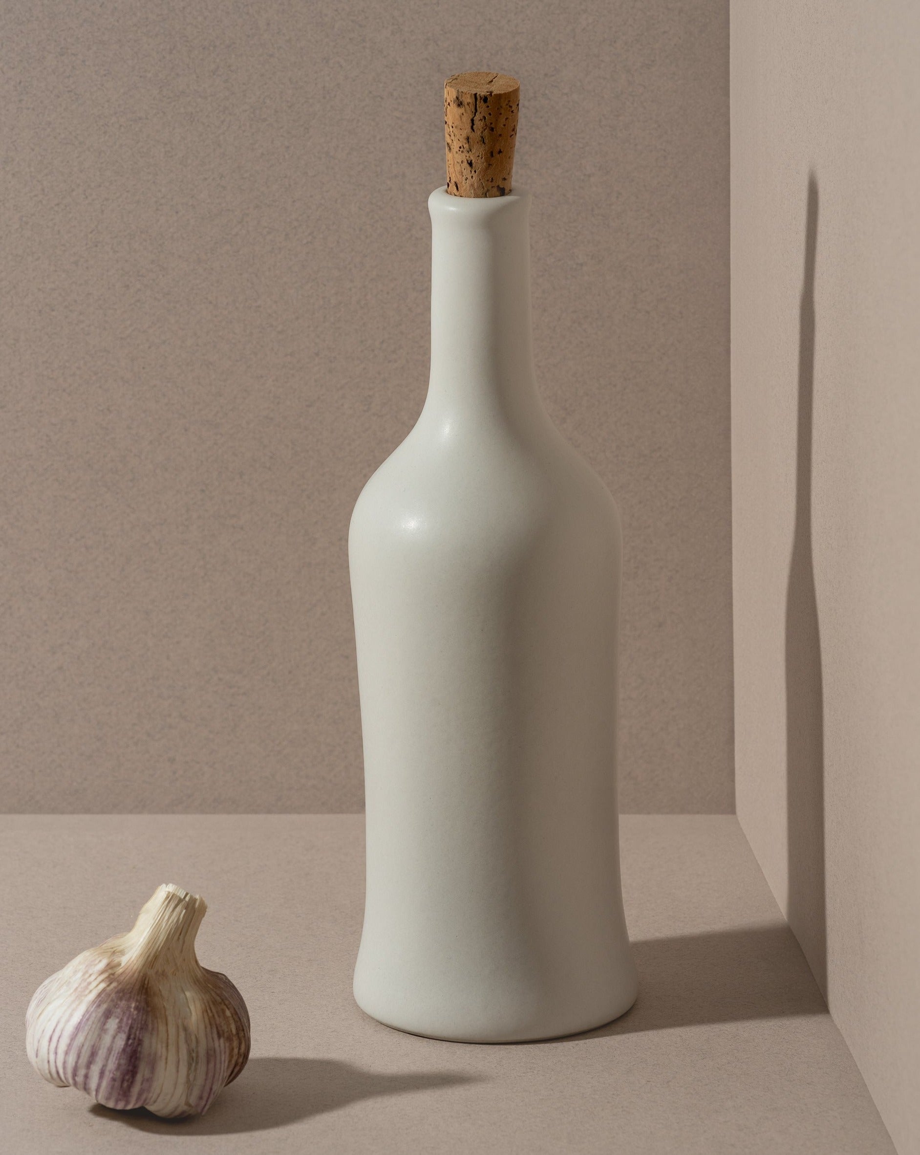 A white ceramic olive oil dispenser with a cork stopper, displayed next to a garlic bulb, on a neutral background.