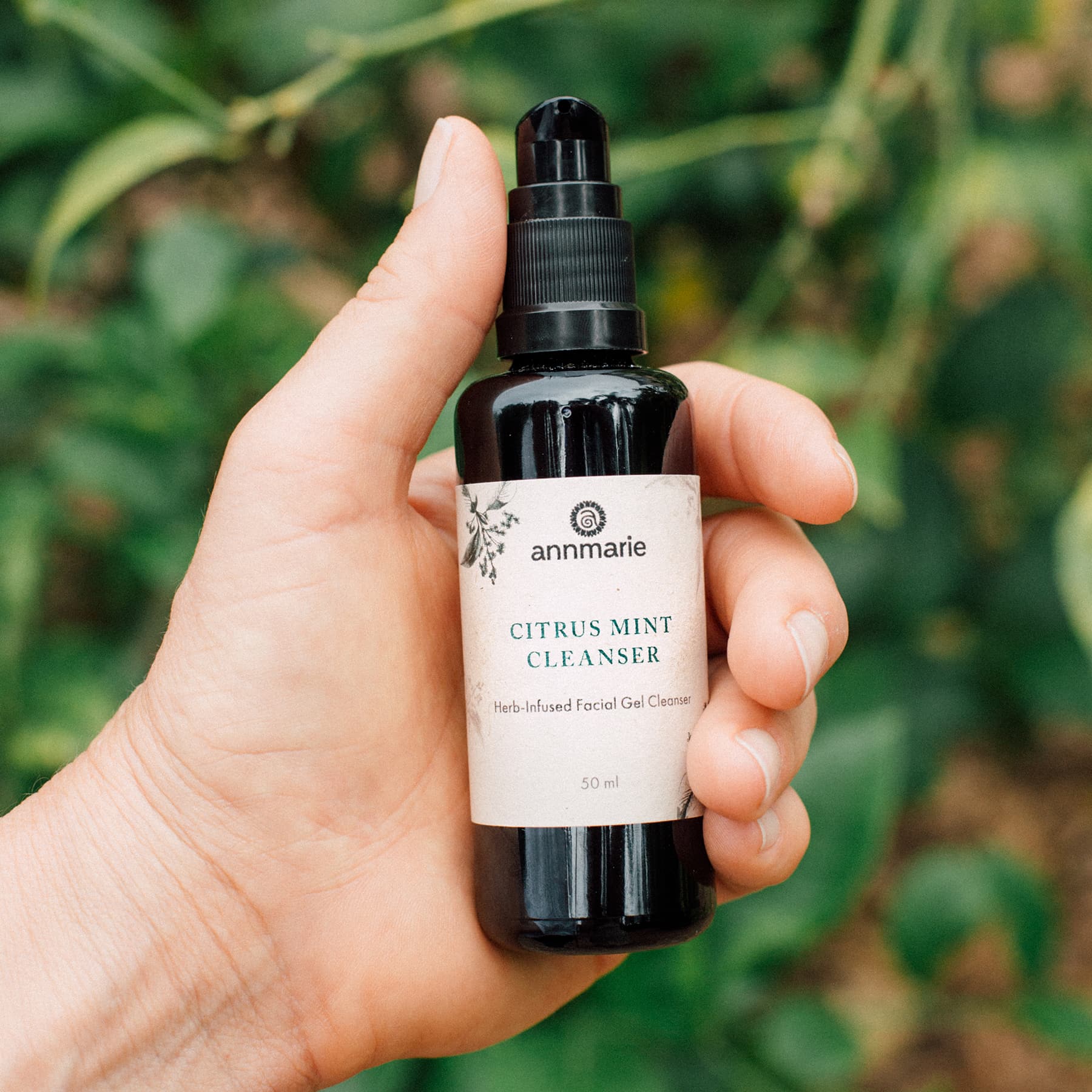 Hand holding a bottle of 'annmarie' Citrus Mint Cleanser against a green leafy background