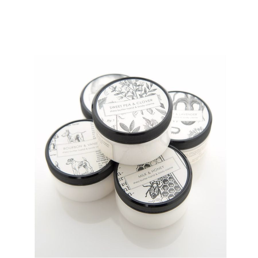 A group of five white jars with black lids are stacked together. Each jar has a decorative label with different text and illustrations, including "Sweet Pea & Clover," "Bourbon & Vanilla," and "Milk & Honey." The background is white and well-lit.
