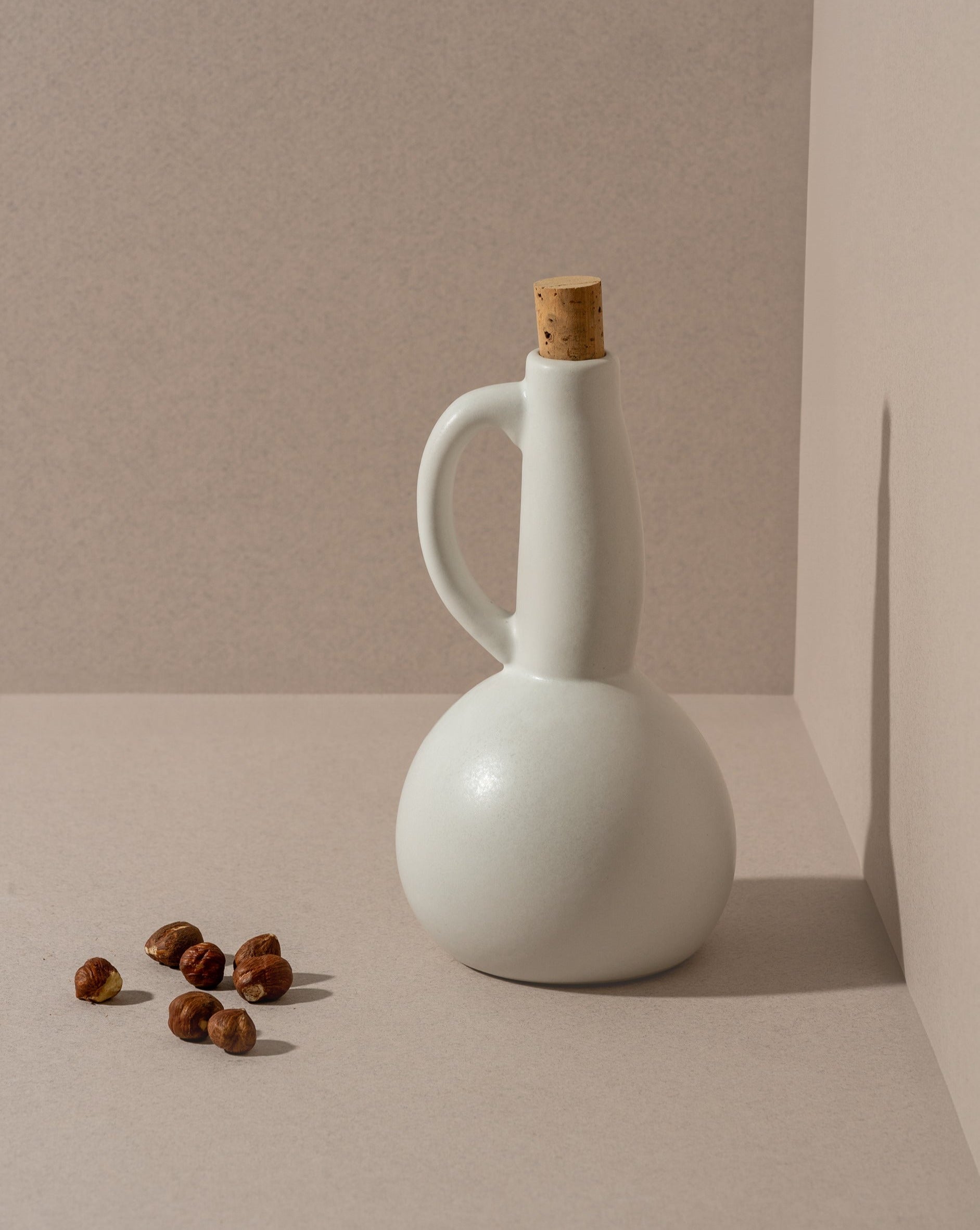 A white ceramic olive oil dispenser with a matte finish, a cork stopper, and a handle, displayed in a corner setting with nuts scattered in front.
