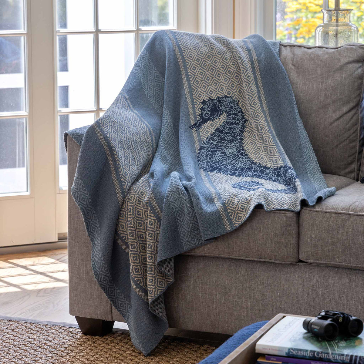 The In2Green Seahorse Square - Cotton Sustainable Knit Throw Blanket in a blue and beige coastal pattern is draped over the arm of a gray sofa in a sunlit living room.