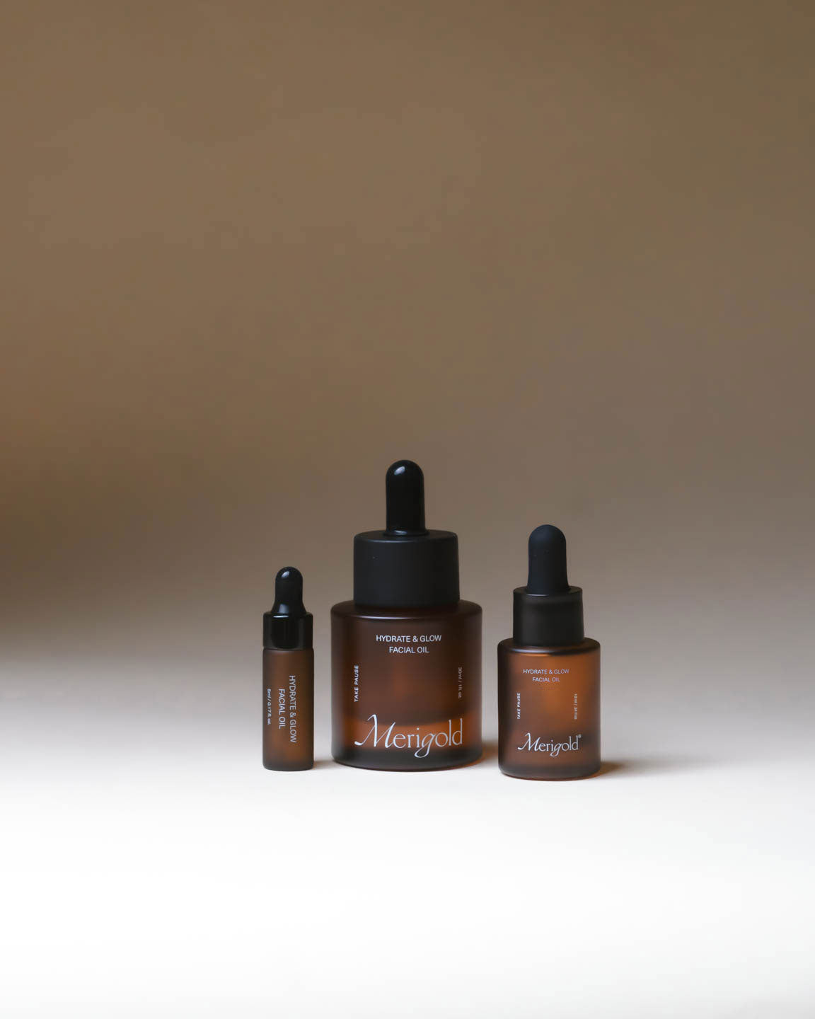 Merigold Hydrate and Glow Facial Oil displayed in amber glass bottles in full size and travel sizes on a neutral background.
