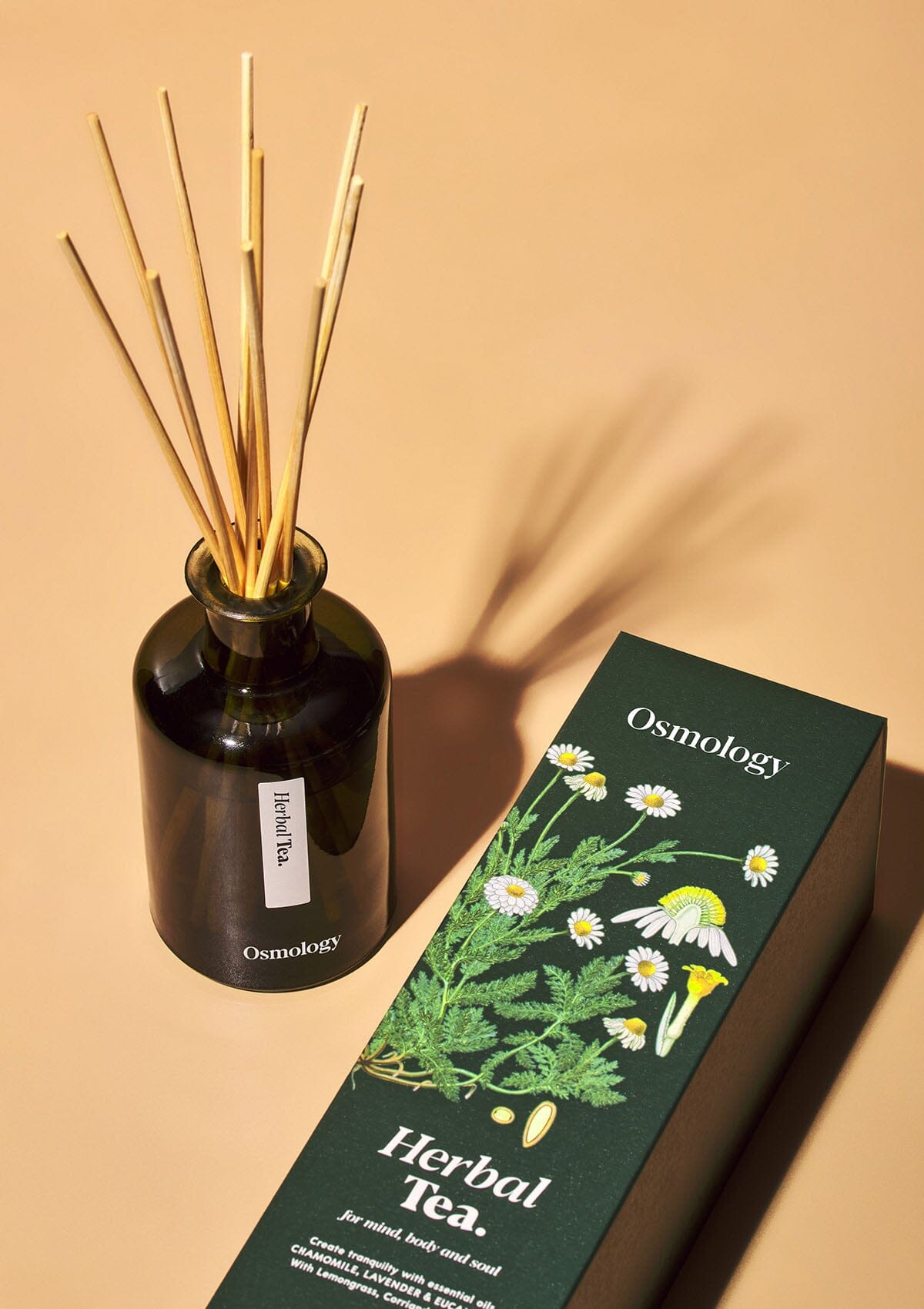Osmology herbal tea reed diffuser in dark green glass next to product packaging on a white background