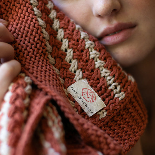 Close-up of a rust red knitted cotton throw with a Pink Lemonade brand label, held by a woman.