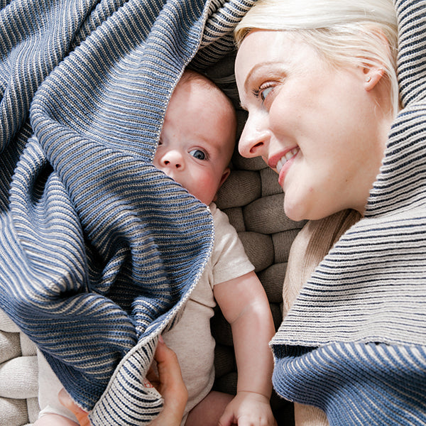 Woman and baby cuddling under a striped marine blue cotton blanket on a cozy knitted mat cushion.