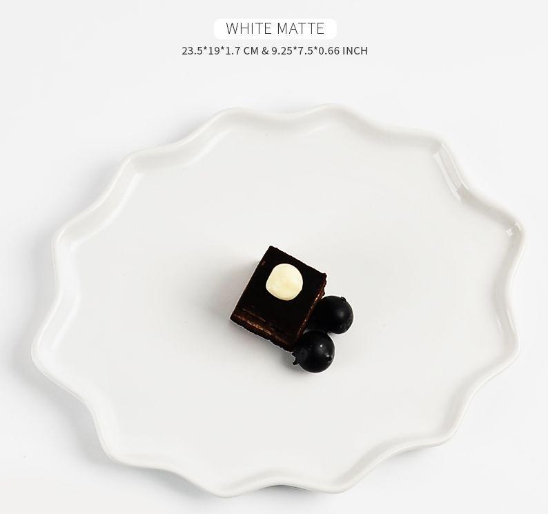 Technical dimension graphic for the BosilunLife Forest Star dinner plate in 'WHITE MATTE' recycled eco-clay. The plate features a unique, scalloped wavy rim and is styled with a small square of chocolate cake topped with cream and black berries. Text at the top details the size: 23.5 by 19 by 1.7 CM (9.25 by 7.5 by 0.66 inches), showcasing its low-profile, organic oval shape.