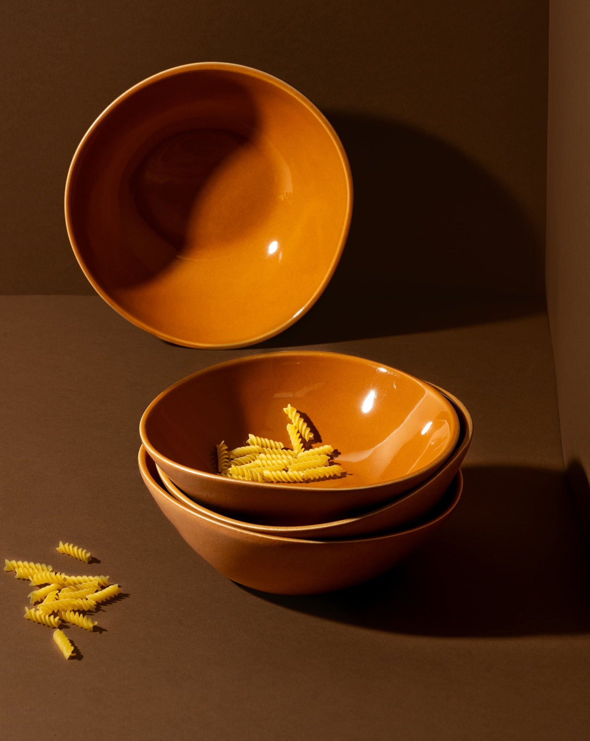 Stack of three handmade, matte black stoneware bowls in terracotta color, with uncooked pasta inside the top bowl; a fourth matching bowl is placed separately beside the stack. Ethically made, artisan-crafted design by Gharyan Stoneware, labeled as dessert plate, soup bowl, and poke bowl. Fair trade, timeless and minimalist aesthetic.