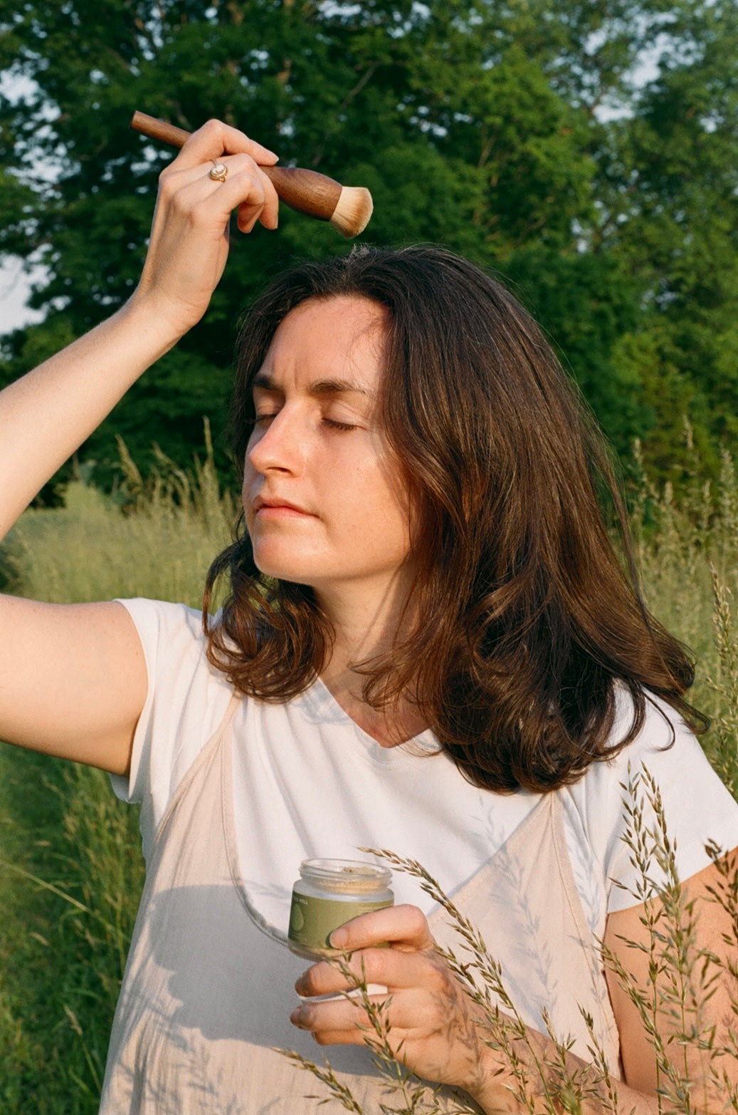 Woman in a field holding a stick and a small container, surrounded by greenery.