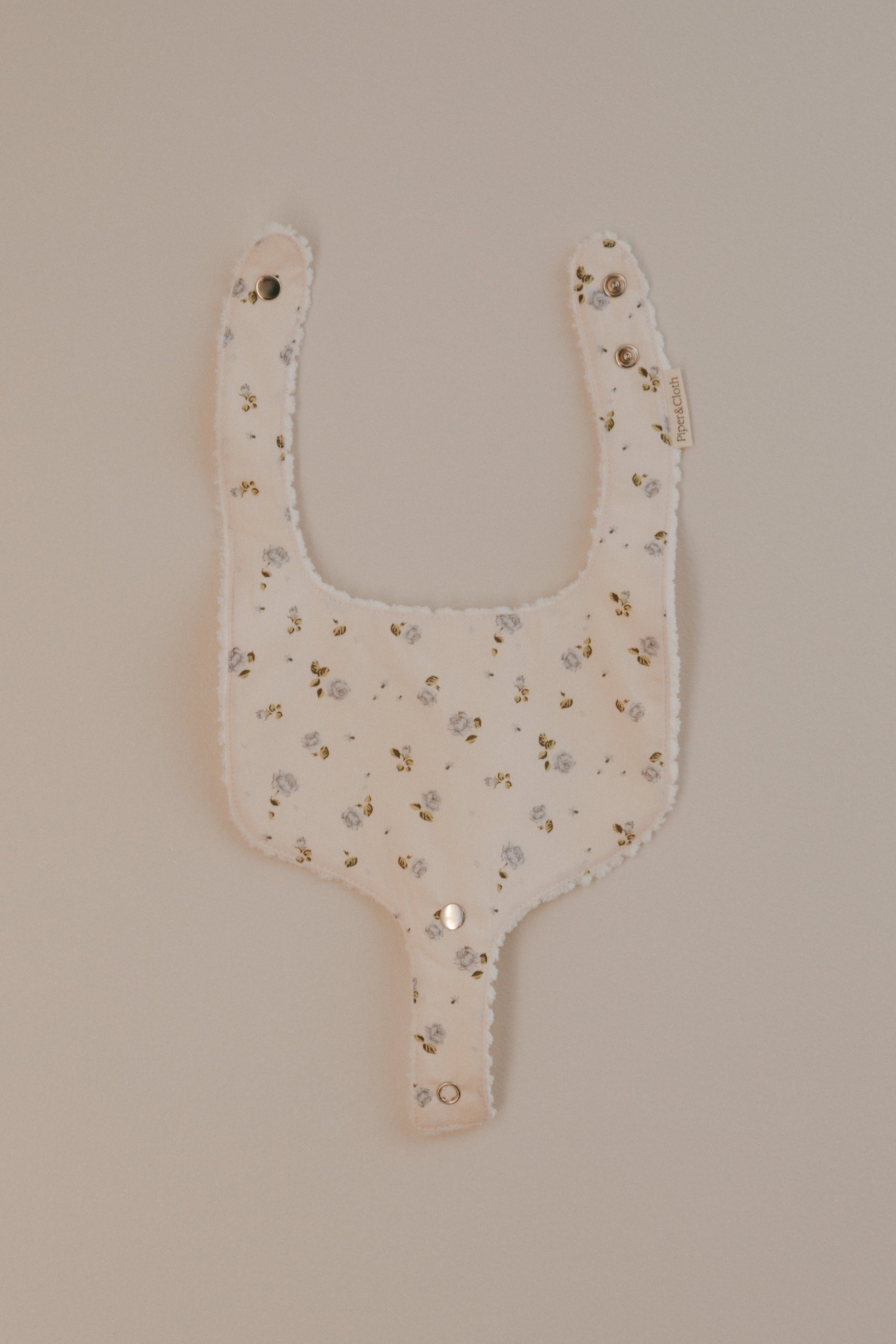 A beige cotton bib with a rosebud print and a pacifier snap, displayed against a neutral background.