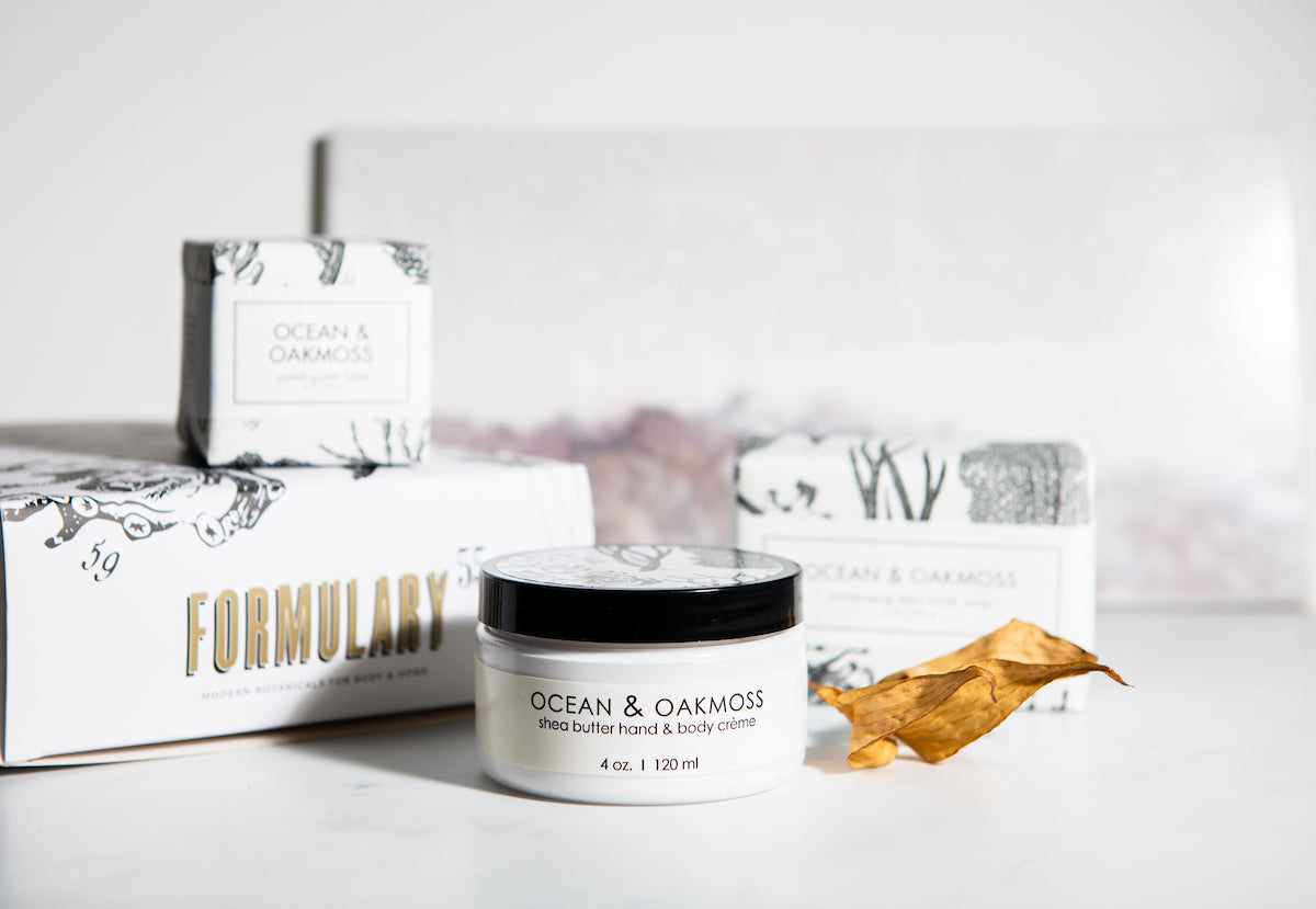 A group of skincare products from Formulary 55 is shown, featuring Ocean & Oakmoss shea butter hand & body creme in a 4 oz. jar in the foreground, and three boxed products with similar branding in the background. A dry leaf lies beside the jar on a white surface.