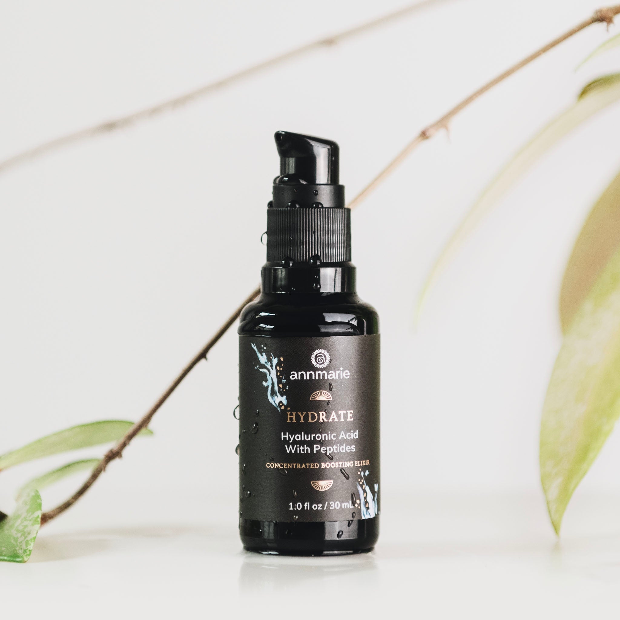 A black pump bottle labeled 'Hydrate' containing a hyaluronic acid with peptides skincare product, placed against a white background with green leaves to the side.