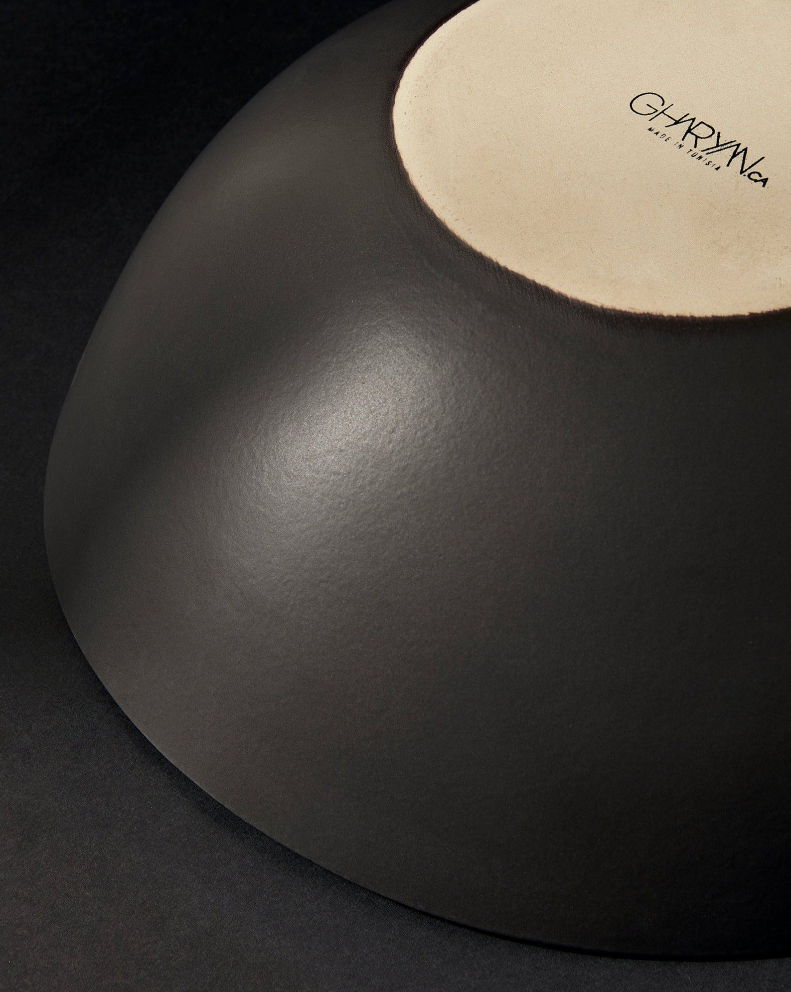 Upside-down black stoneware serving bowl with the 'GHARYAN' logo visible, partially shown against a black background.