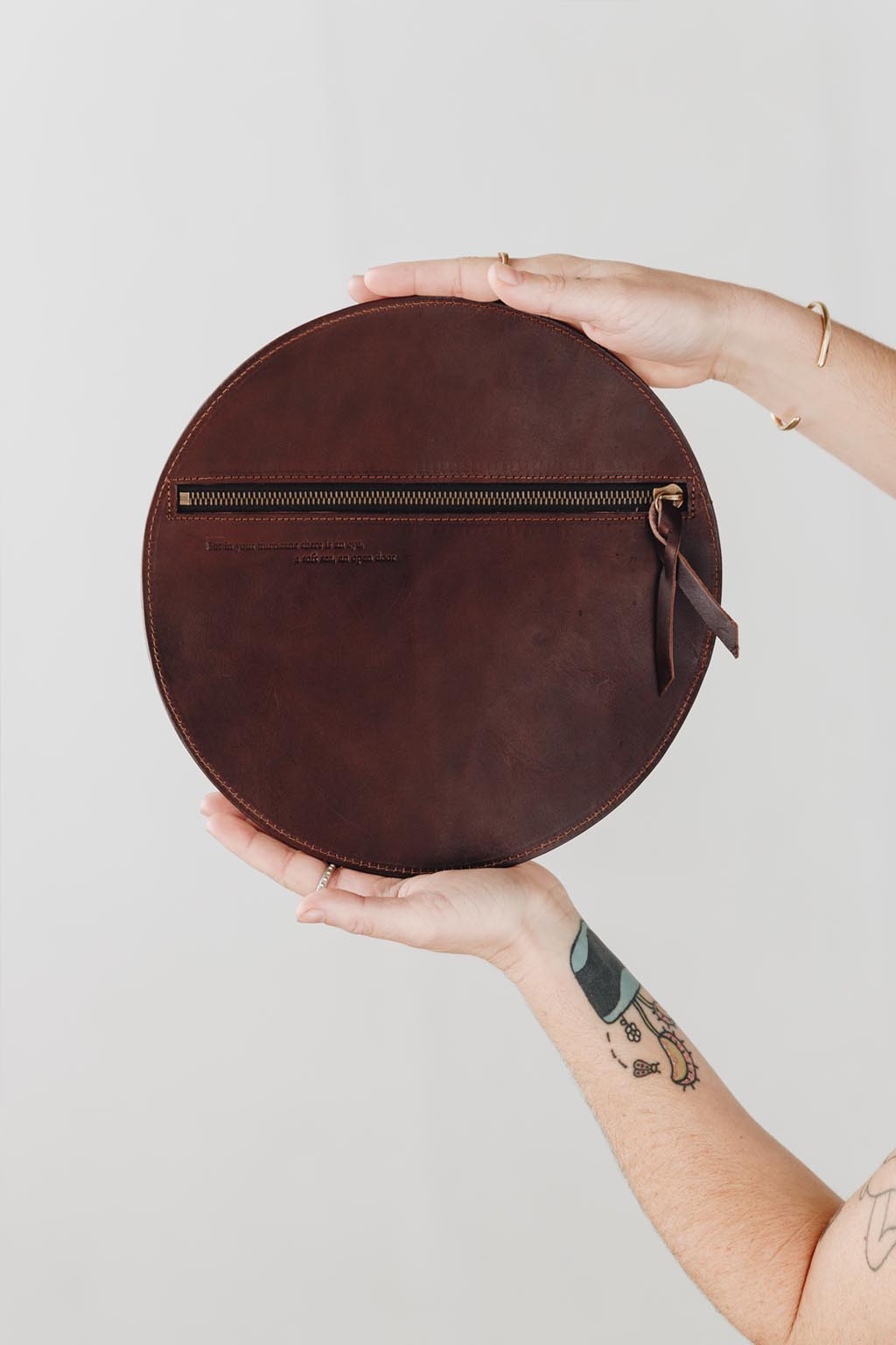 A woman's arms are visible, holding a circular dark brown leather clutch with the reverse side and zipper visible. The zipper includes a matching zipper pull.