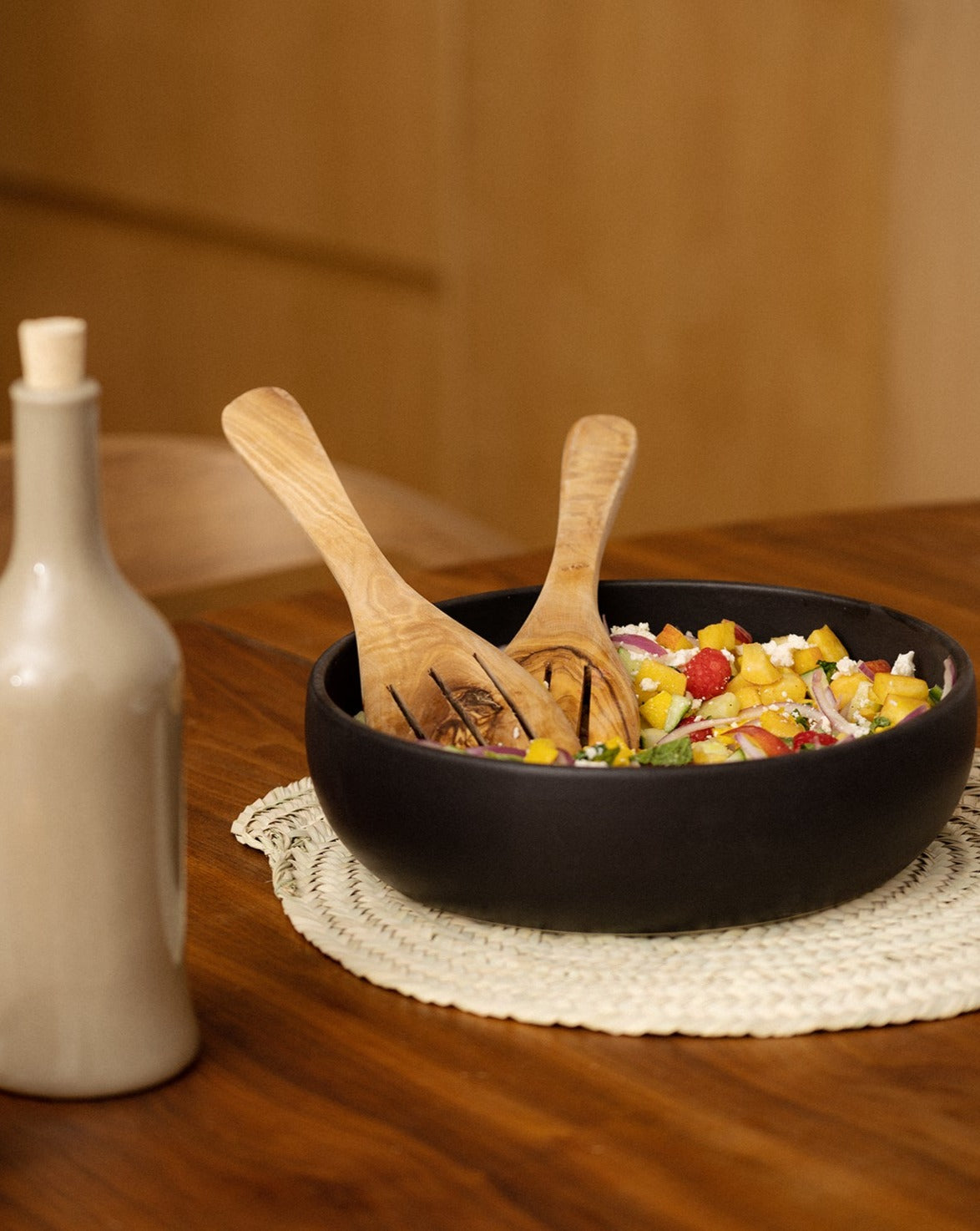 A black stoneware shallow serving bowl filled with salad, featuring a wooden serving spoon resting inside. Beside the bowl is a white olive oil dispenser, all set on a wooden dining table.