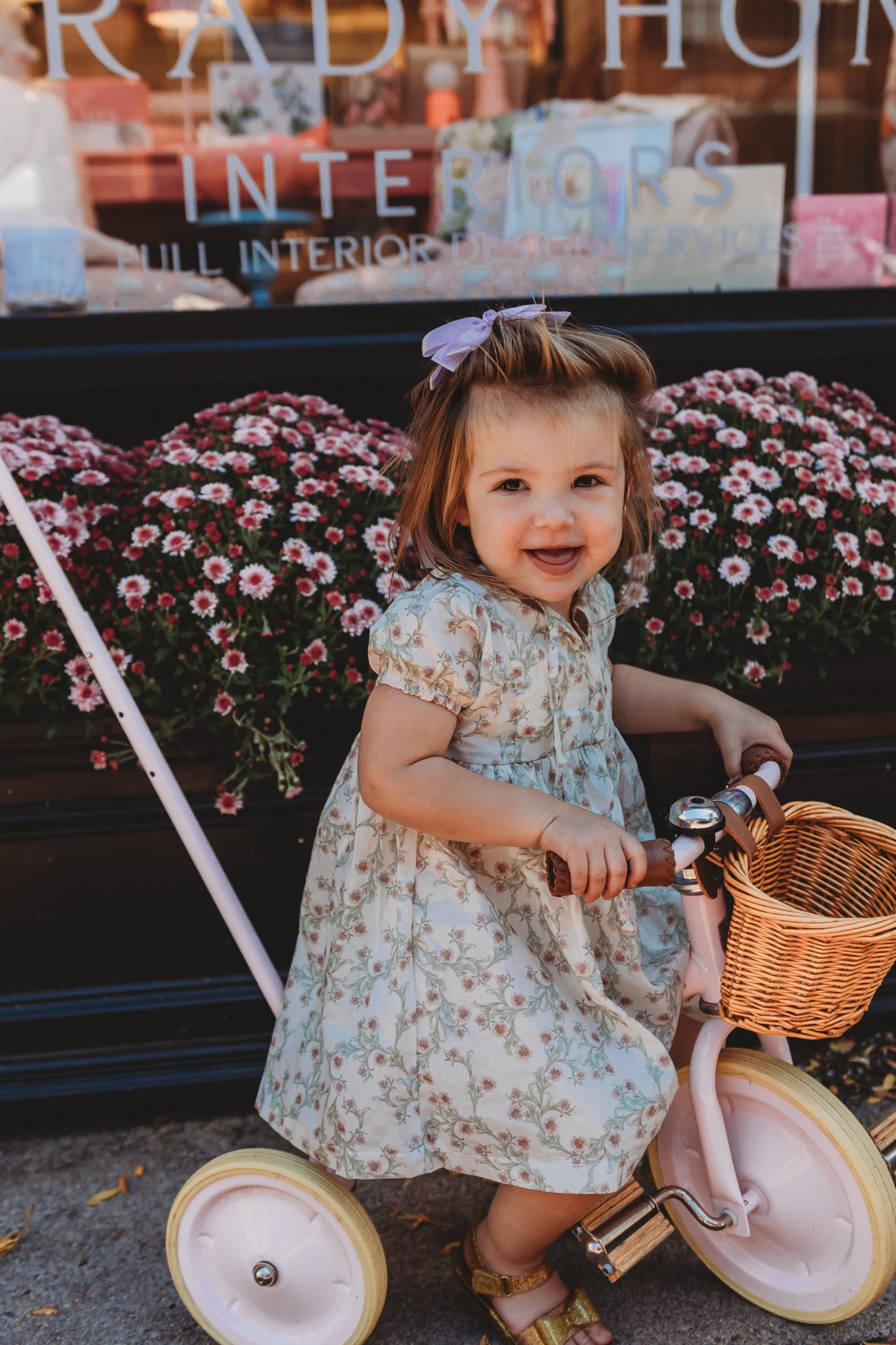 Young girl riding a tricycle with a basket in front of a store with flowers in the background