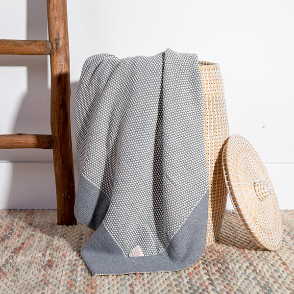 Light grey textured cotton throw draped over a woven cork basket on a patterned rug next to a brown ladder.