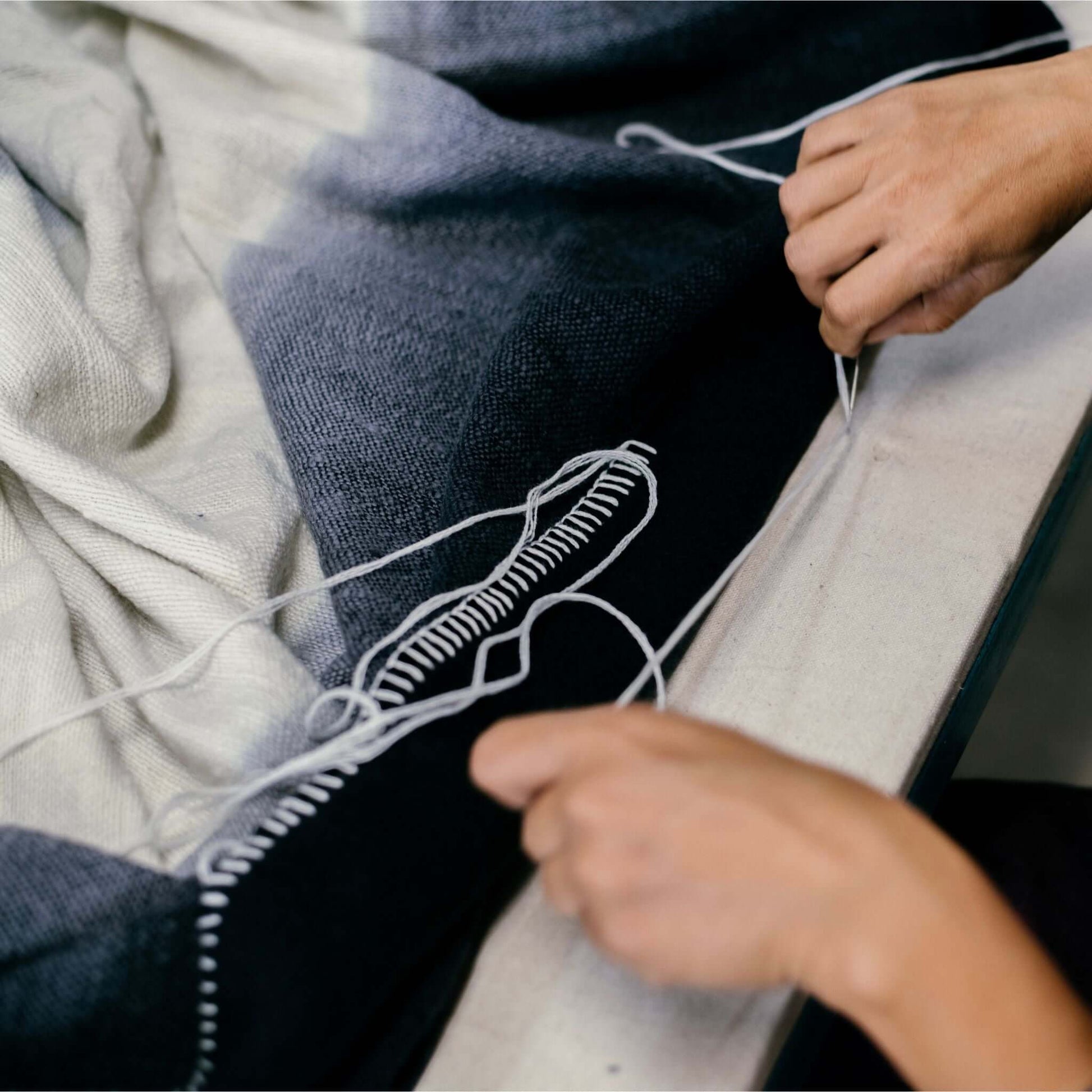 Person hand stitching the hem of the Ink Black White Ombre Textured Throw In Hand Spun Merino And Hand Stitched Hem.