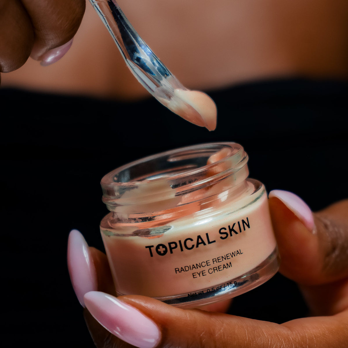 A close-up of a person holding an open jar of TOPICAL SKIN Radiance Renewal Eye Cream while applying cream from a small metal spatula to their eyelids.