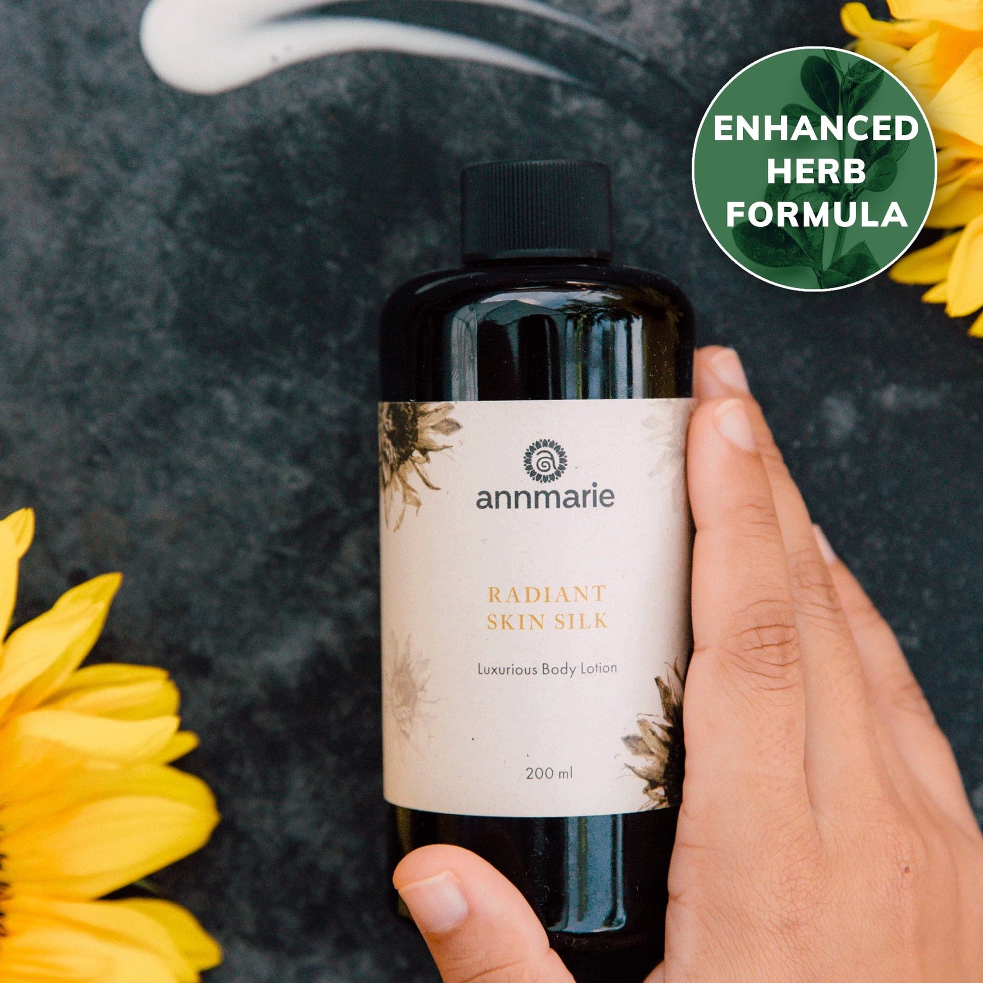 Hand holding a bottle of Annmarie Radiant Skin Silk lotion with sunflowers on a dark background