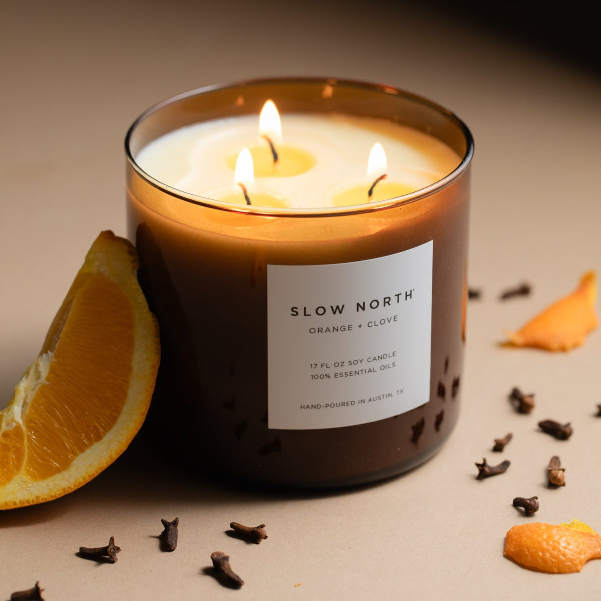 Candle with 'Slow North' label surrounded by orange slices and cloves on a beige background