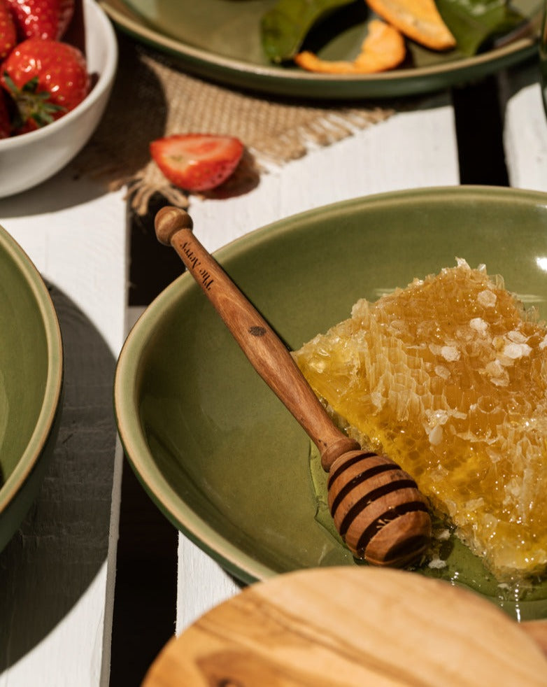 Green bowl filled with honey and an olive wood honey spoon, placed beside a plate with strawberries and food.