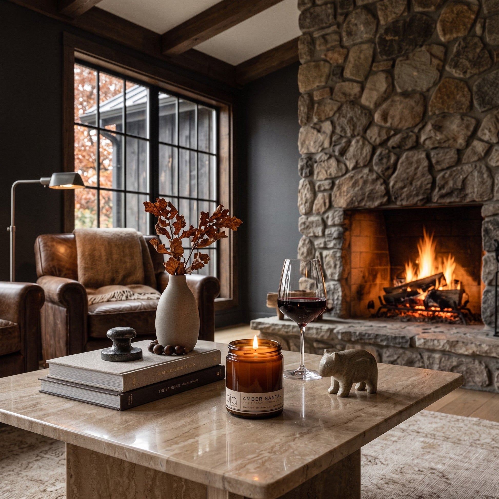 A cozy living room scene with a stone fireplace burning brightly, a leather armchair by a window, and a marble coffee table holding a lit candle, books, a vase with dried branches, a glass of red wine, and a small bear figurine.