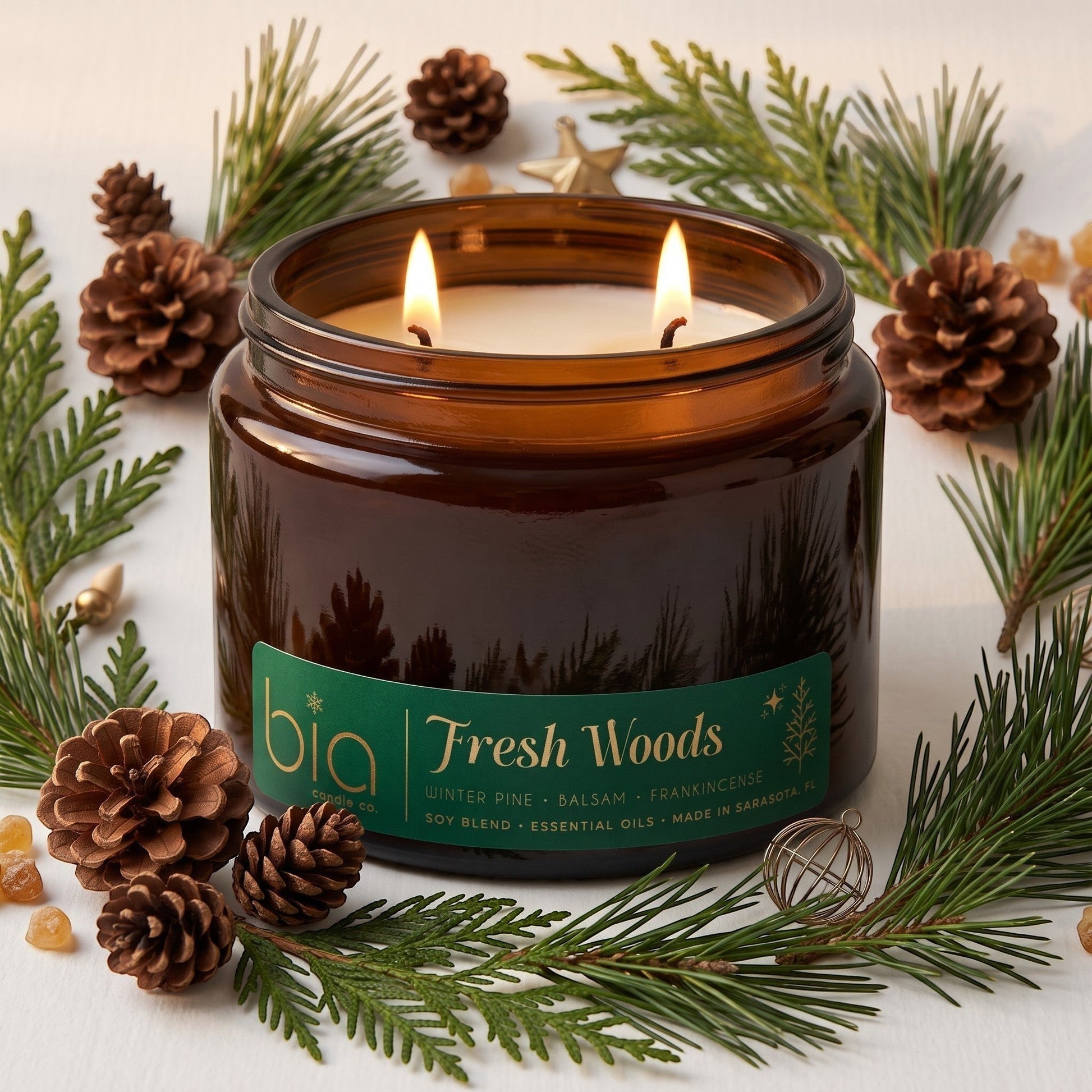 A grande lit amber glass jar candle with two flames, labeled 'bia Fresh Woods', surrounded by pine branches, pinecones, and small gold decorative accents on a light surface.
