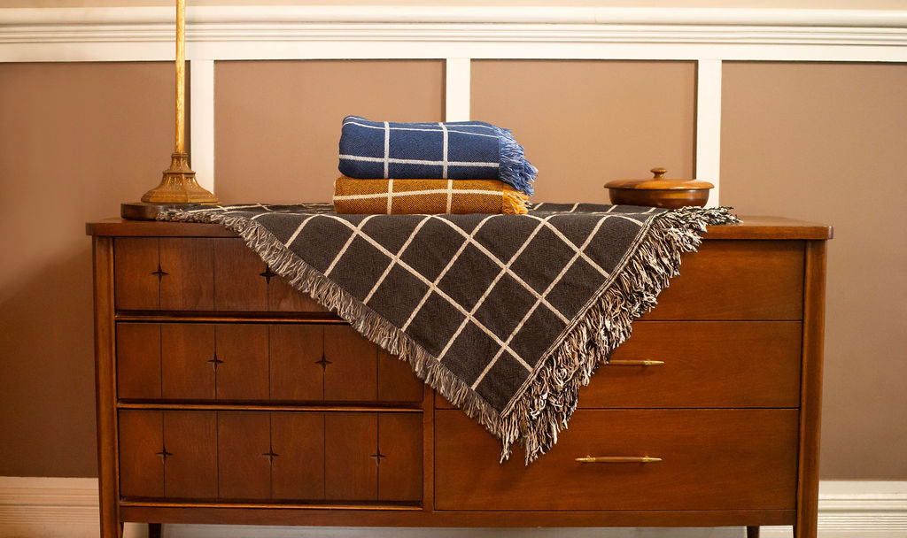 Greer Nocturne grid blanket draped over a vintage wood dresser with blue and goldenrod colorways stacked above