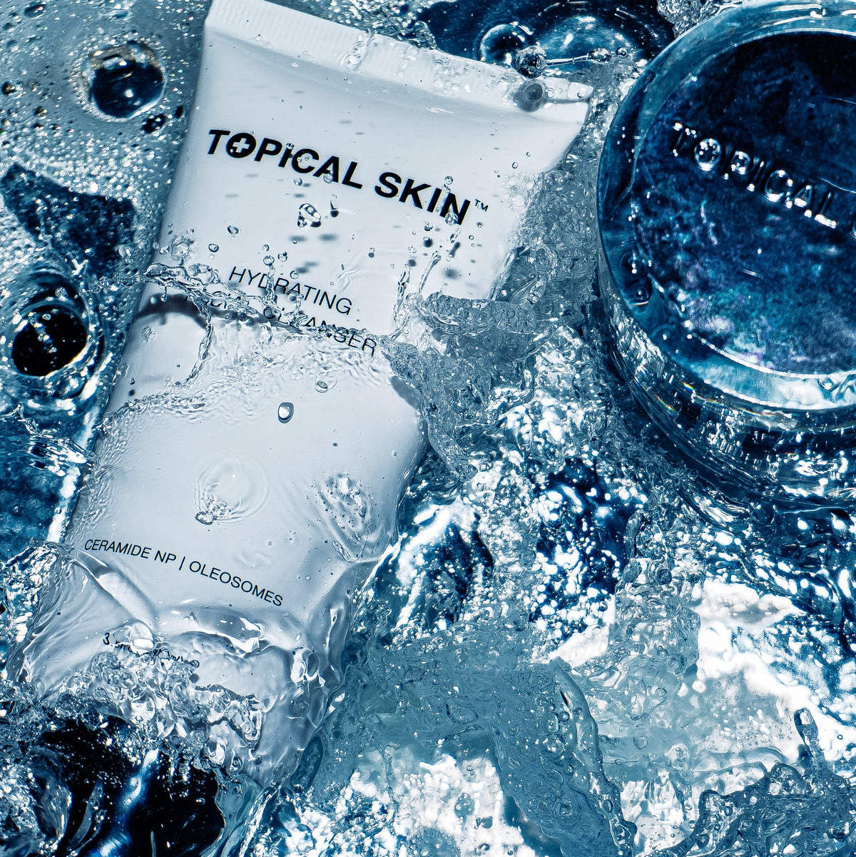 A white tube of TOPICAL SKIN Hydrating Cleanser surrounded by splashing water and ice, with a matching round blue jar partially visible at the top right.