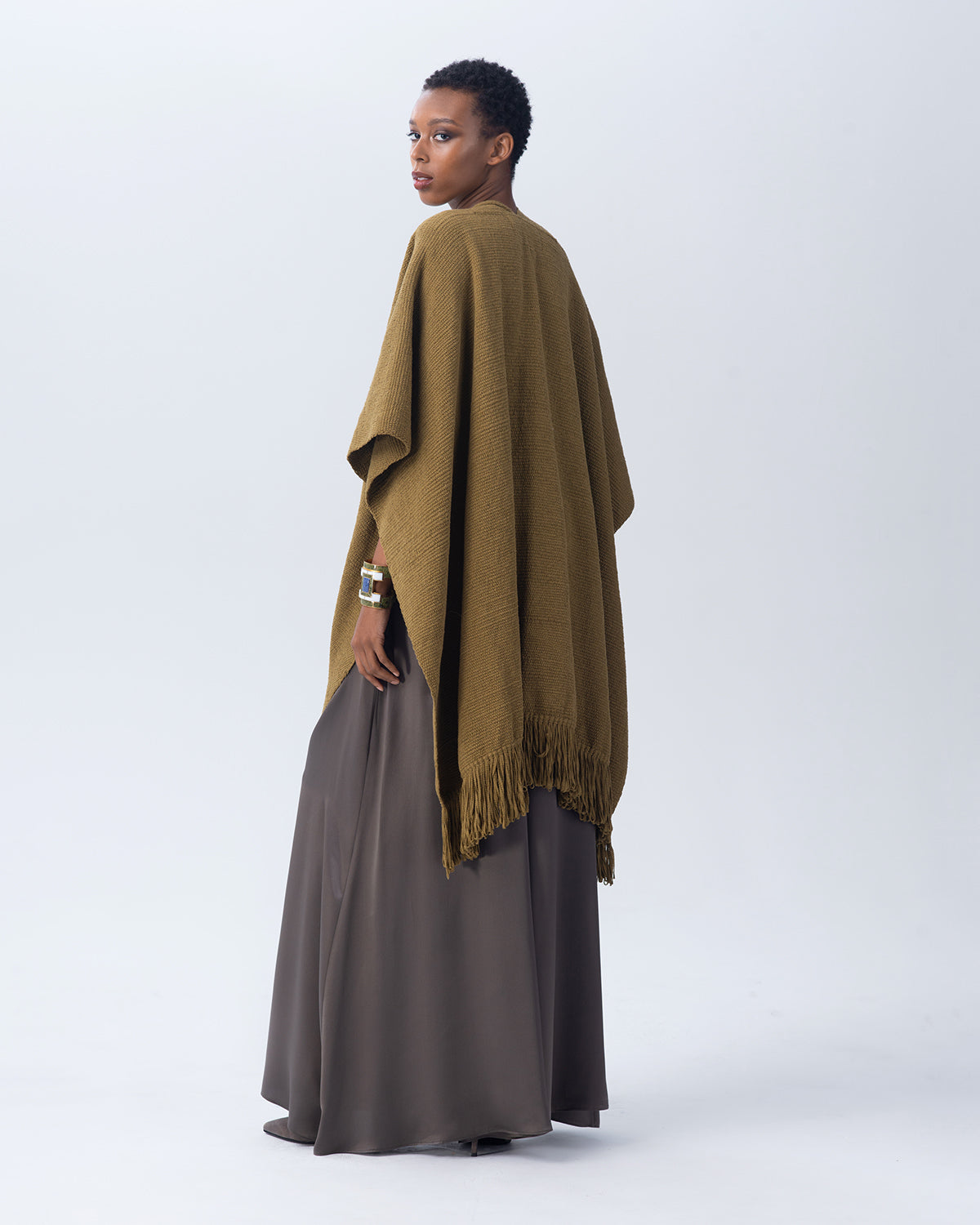 Model wearing an olive Cotton Duna Fringe Duster poncho, standing with back to camera.