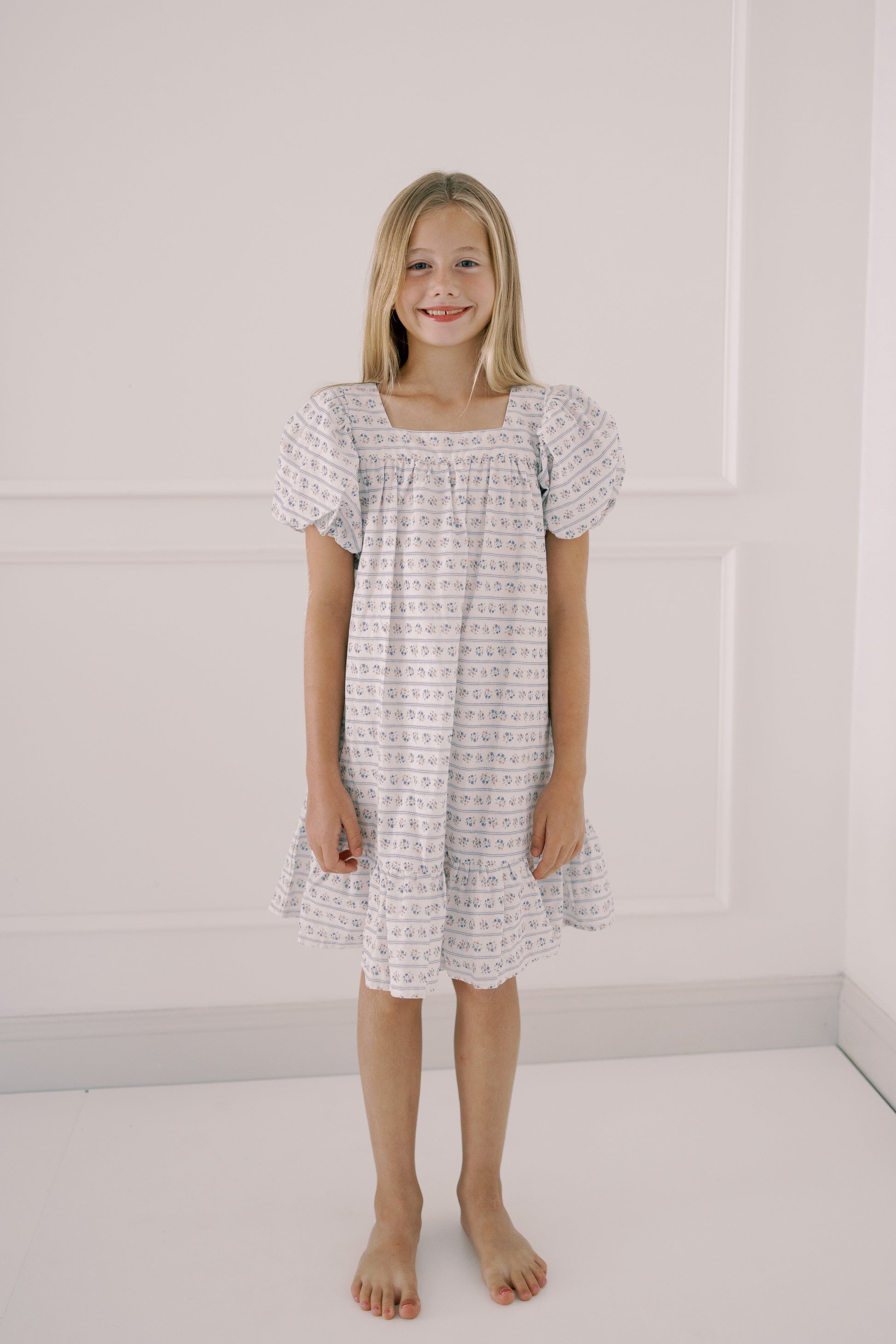 Young girl wearing a patterned dress standing against a white wall.