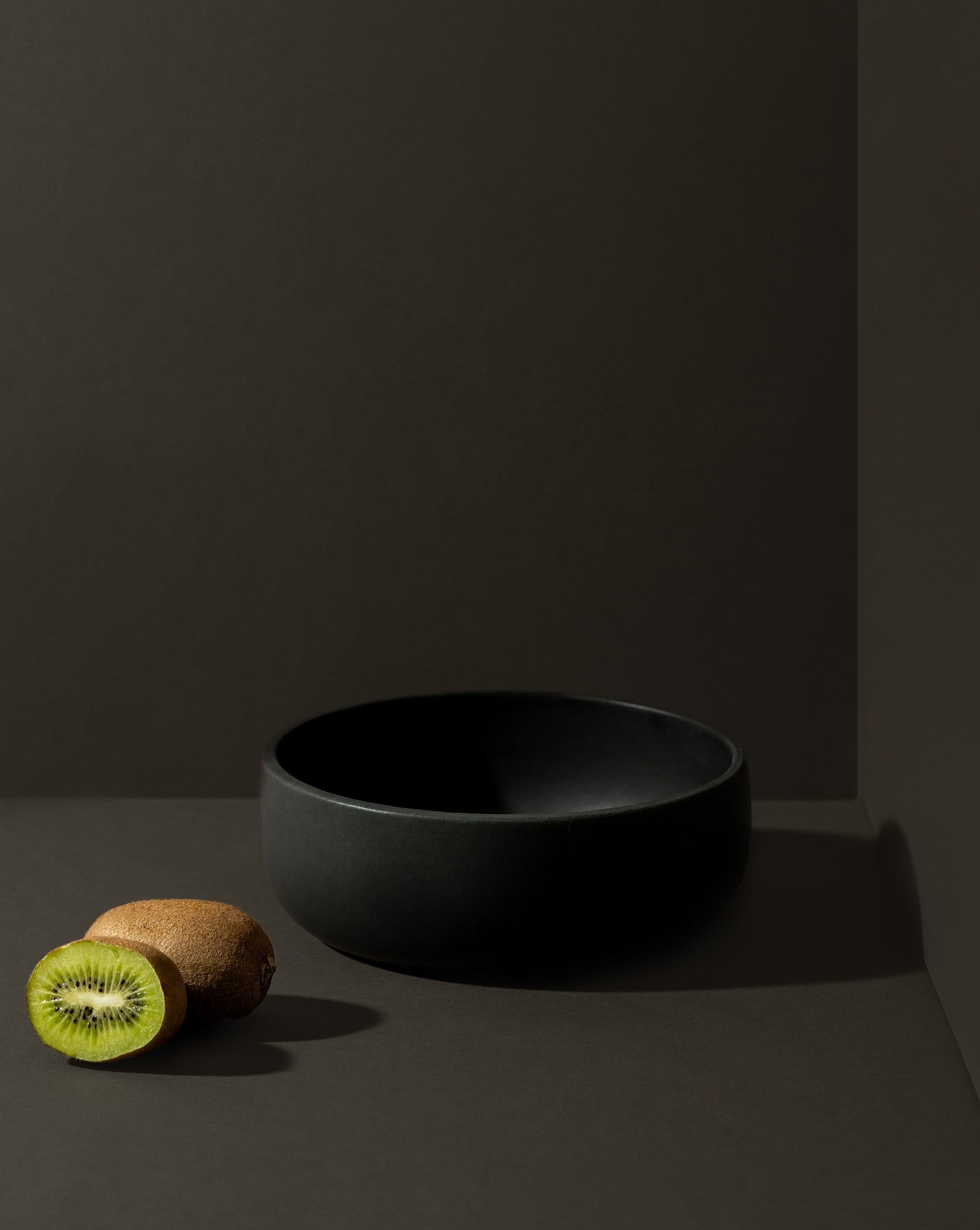A matte black stoneware serving bowl displayed against a dark background, with a sliced kiwi placed beside it to add a natural touch.