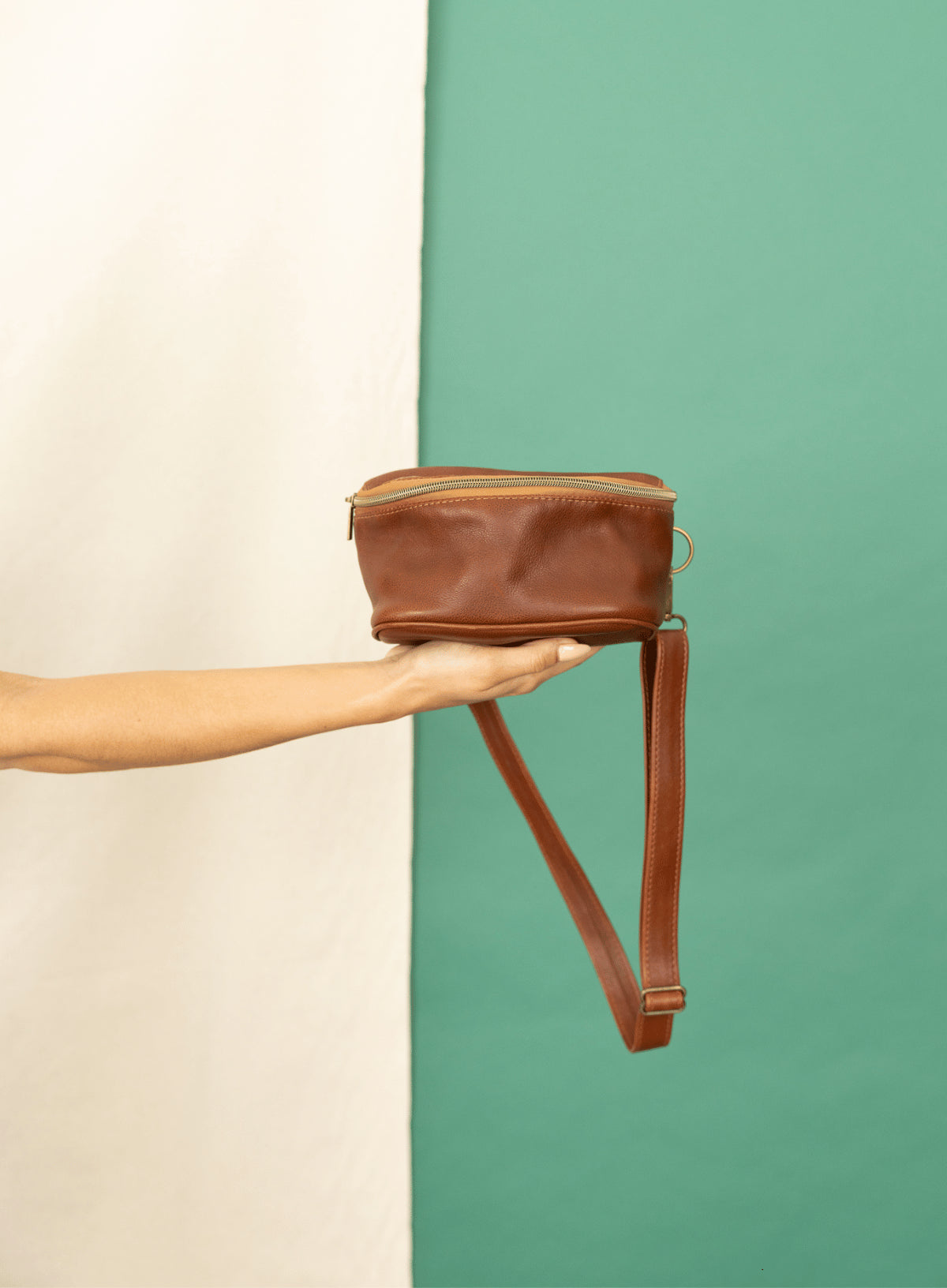 Hip bag in mahogany from the fall/winter collection held by model leaning on a table.