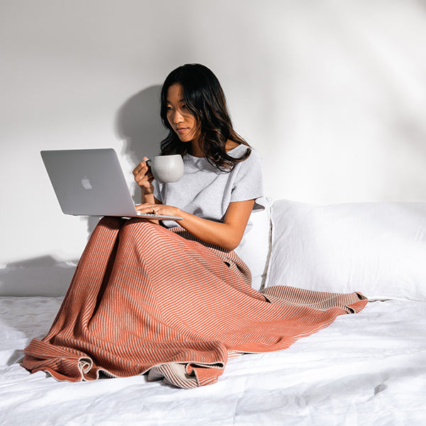 Woman sitting on a bed and working on an Apple laptop and holding a cup of coffee while wrapped in a orange and rust cotton throw.