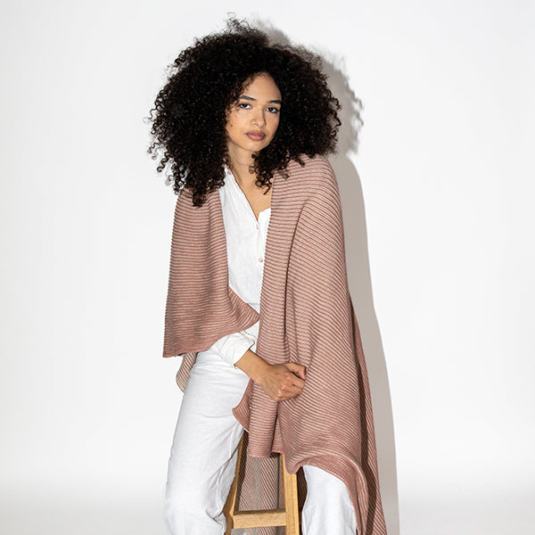 Woman wearing a pink and brown throw over a white outfit while sitting on a wooden stool. 