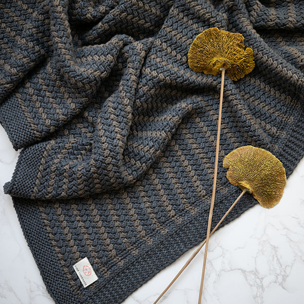 Knitted cotton blanket with a textured dark grey and blue pattern on a white surface, accompanied by dried leaves.