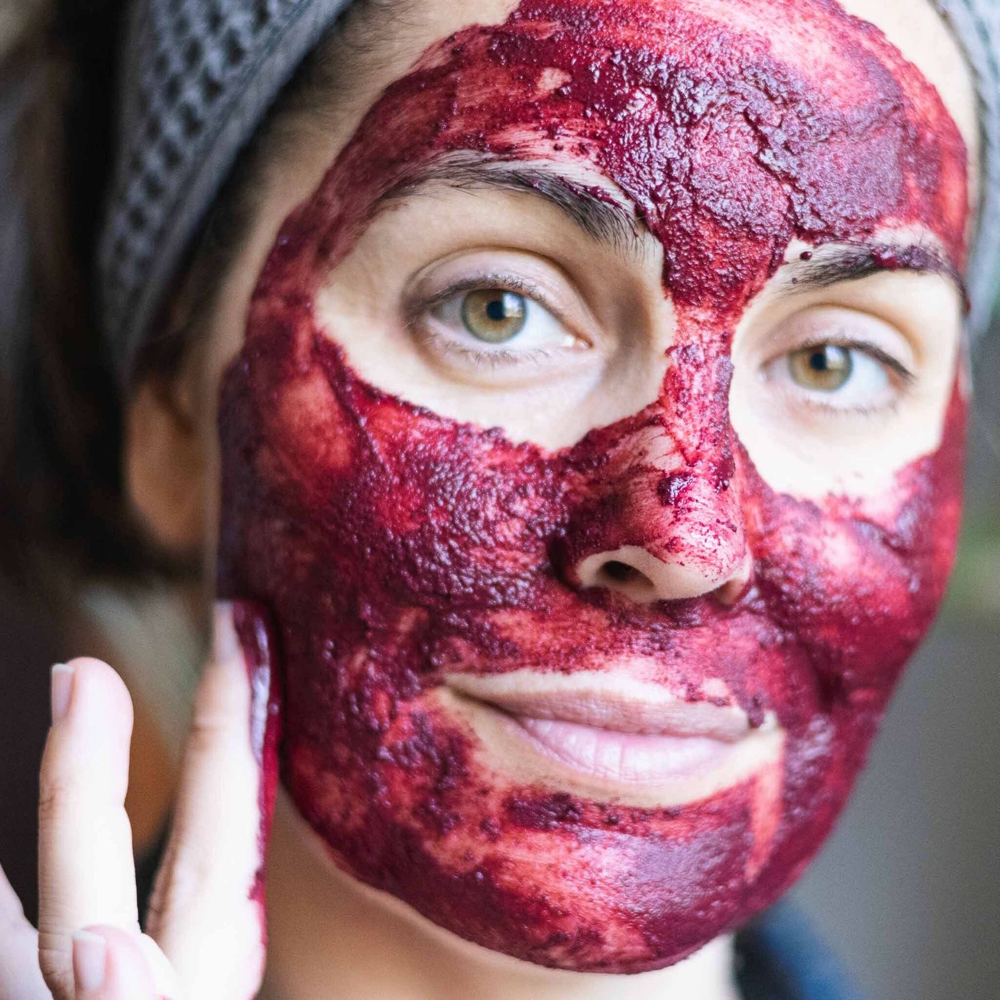 Person applying a deep magenta Annmarie Radiant-C Mask & Polish treatment to the face, showing the textured botanical exfoliating mask during use.