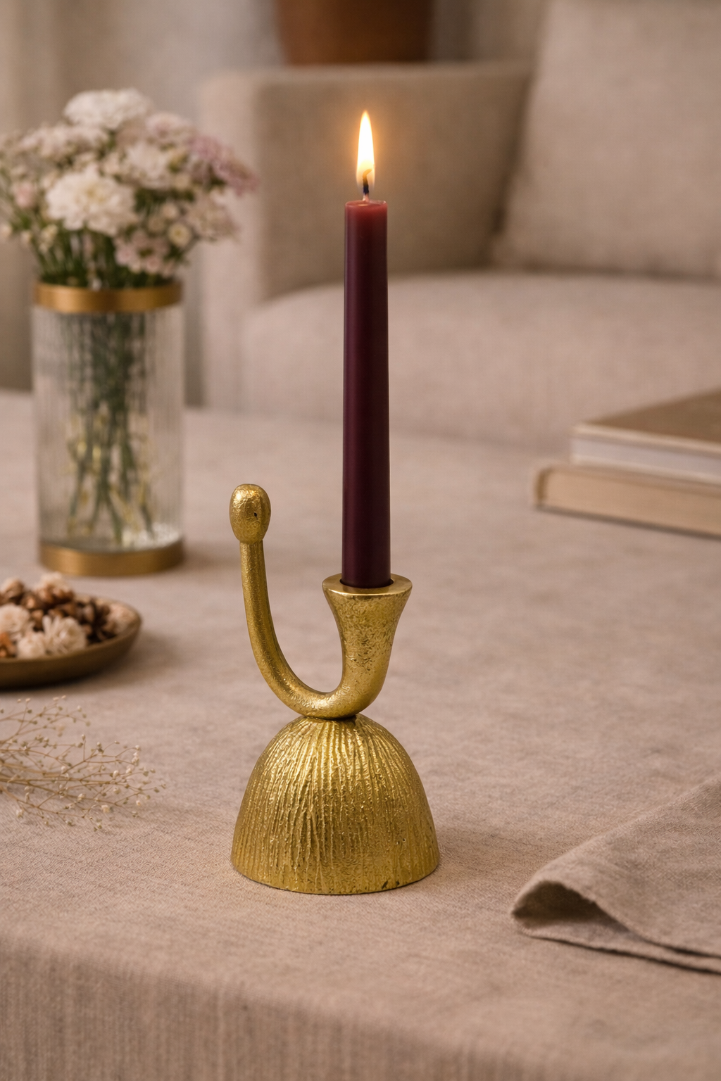 Gold candle holder with a lit purple candle on a table with flowers and books in the background
