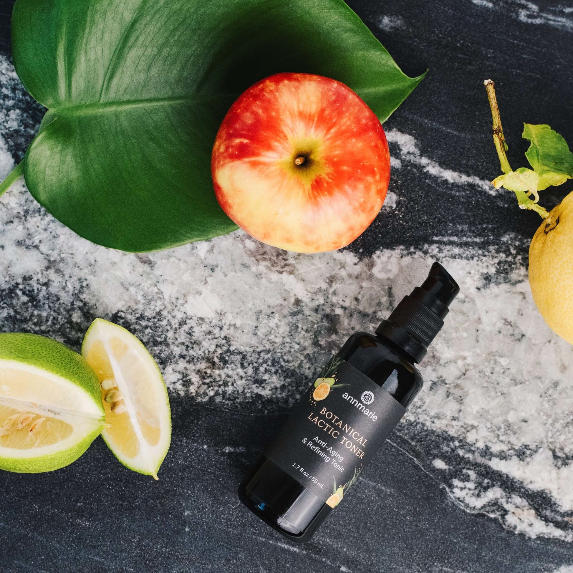 Bottle of Botanical Lactic Toner with an apple, lemon, and lime on a dark surface