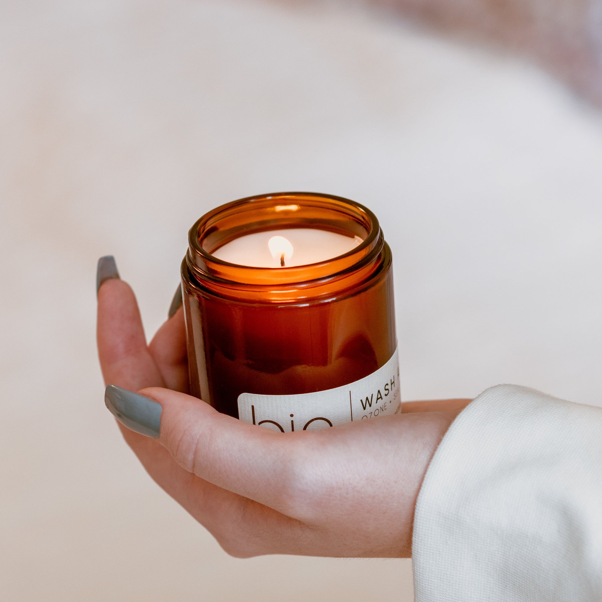 A lit amber glass jar candle held in a person's hand. The candle has a white label on brown jar
