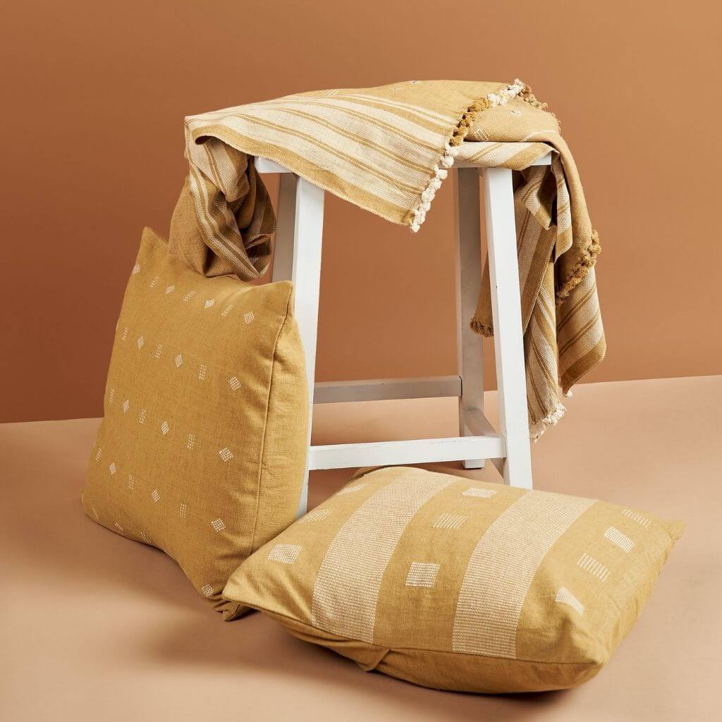 Nira Ochre Hand Spun Organic Cotton Handloom Pillow displayed with a striped throw on a white stool.