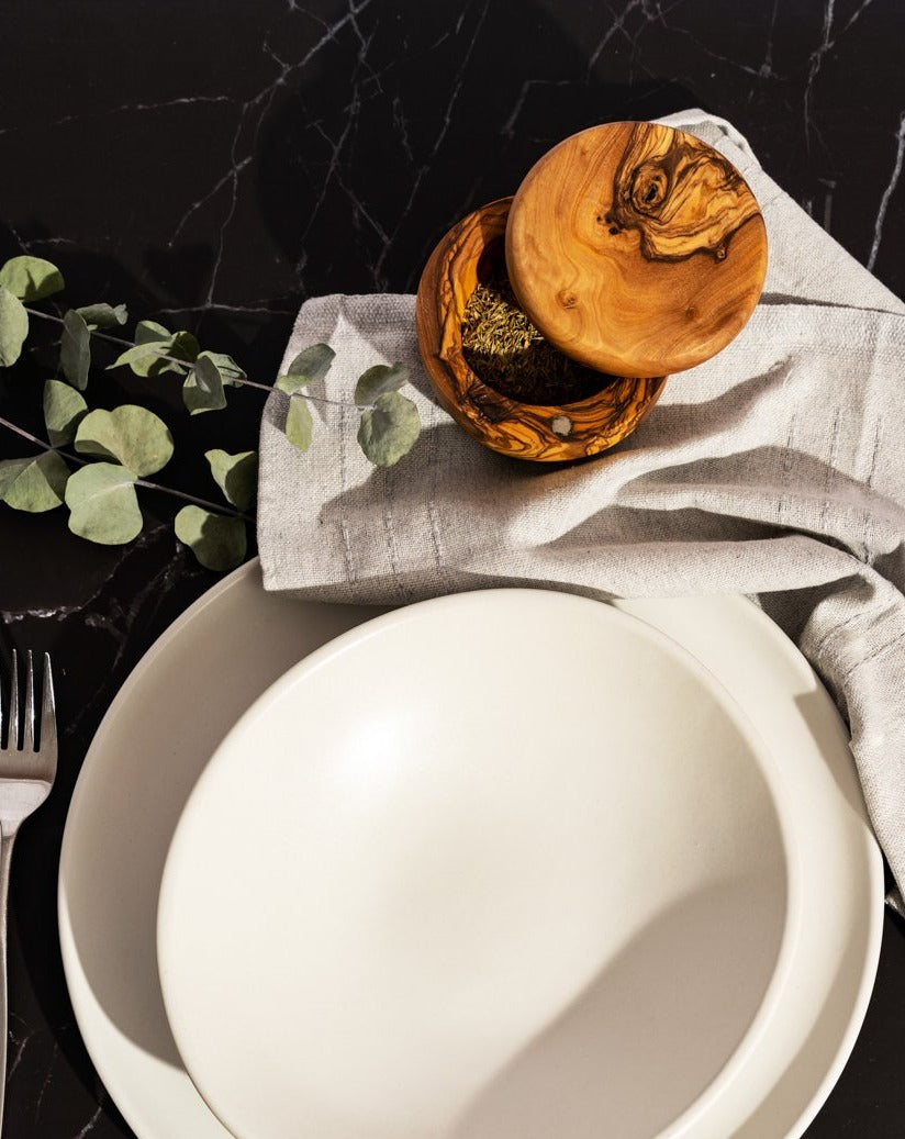 Round olive wood salt and spice cellar placed beside a white plate with a fork, and a decorative stem with leaves.