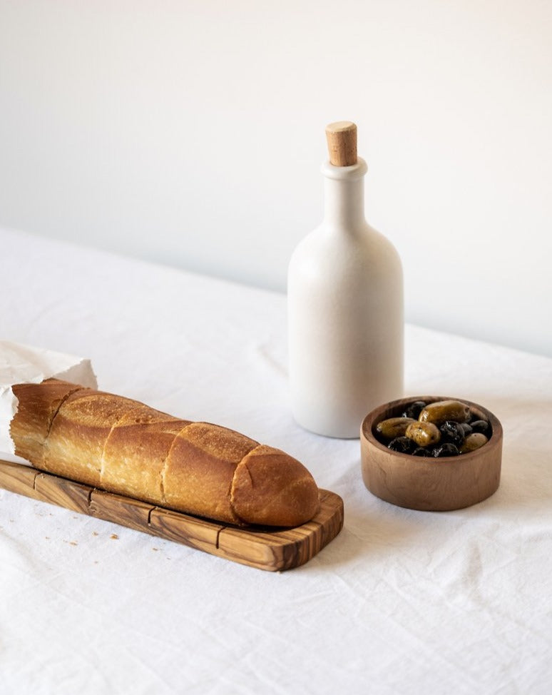 An olivewood sling bread board with visible natural cracks and imperfections, displayed on a dark background. Its unique shape and grain patterns highlight the rustic, handmade quality of the piece.