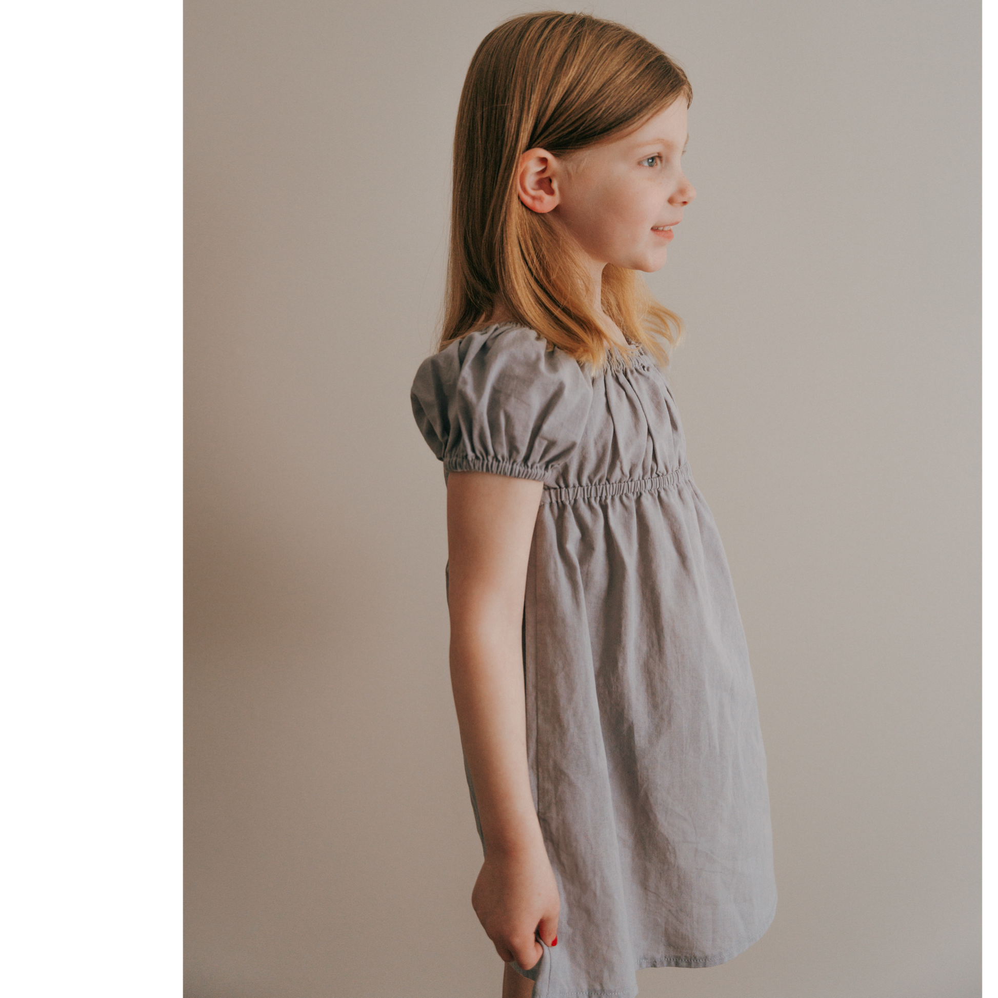 A young girl wearing a short sleeve muted purple dress with cap sleeves, standing against a neutral background.