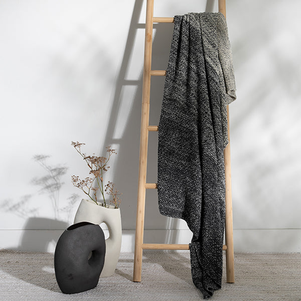 Decorative items including a vase, a plant, and a gray textured throw blanket on a wooden ladder against a white wall.