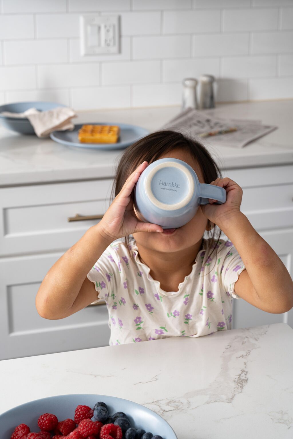 Child drinking from a blue Hamkke Home ceramic mug in a bright kitchen with breakfast plates and berries.