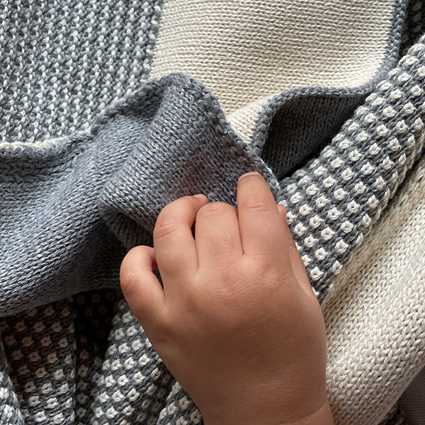 Close-up of a baby's hand holding a textured gray and white cotton throw.