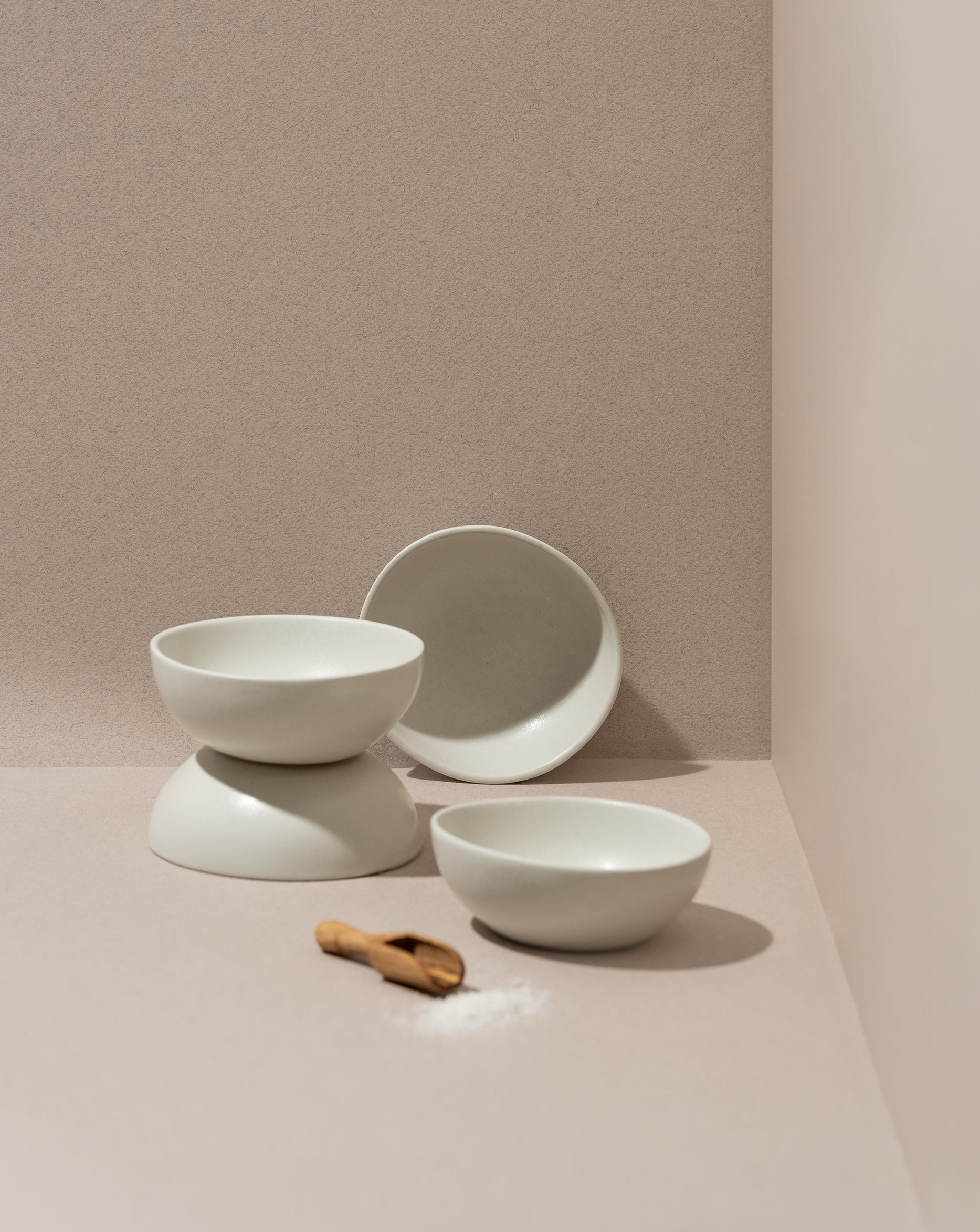 A set of white stoneware condiment bowls with a matte finish, displayed against a neutral background. One bowl is upright, while the others are stacked behind it, and there is a small wooden spoon in front.